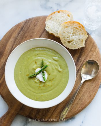 A white bowl of creamy green Asparagus Soup served on a round wood serving board with slices of bread.