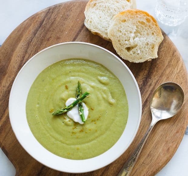 A white bowl of creamy green Asparagus Soup served on a round wood serving board with slices of bread.