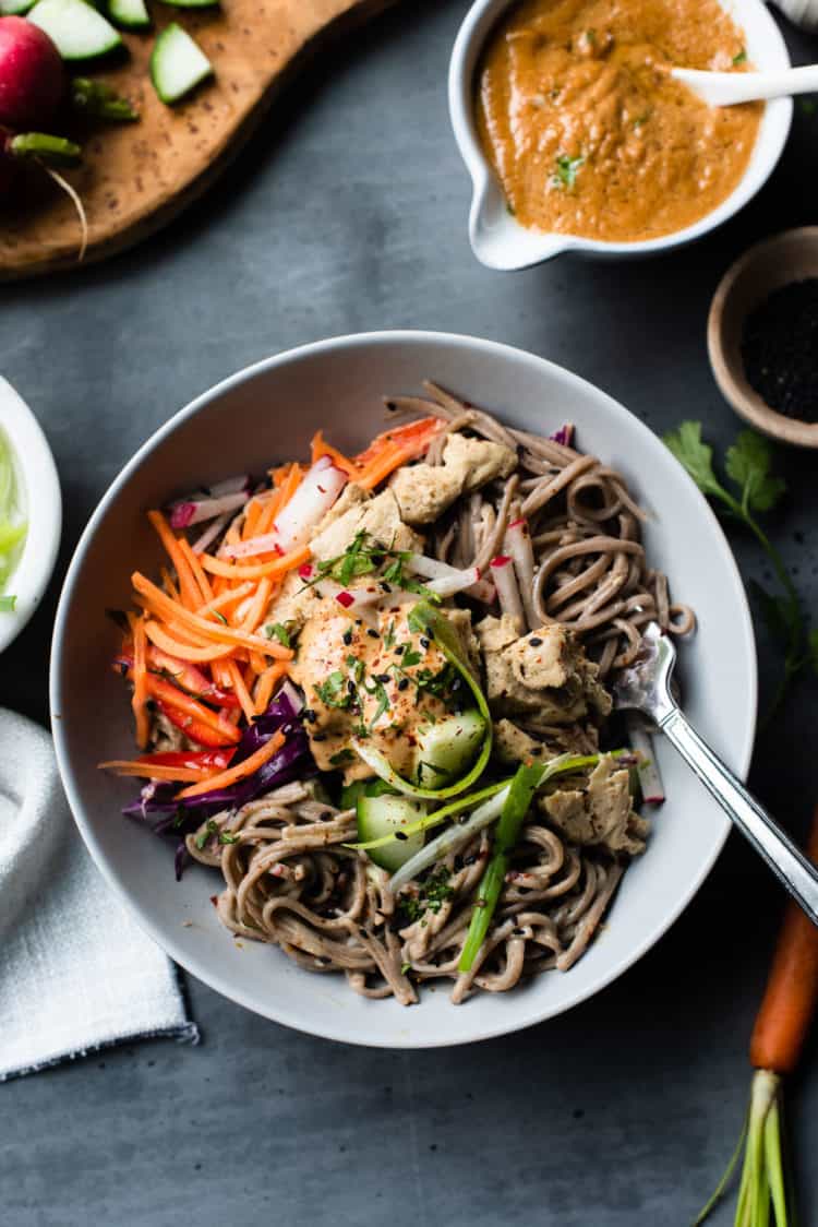 A bowl of soba noodles with chicken, carrots, red cabbage, cucumber, and other vegetables on top.