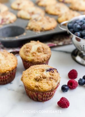 Freshly baked mixed berry muffins with summer berries surrounding them on a countertop.