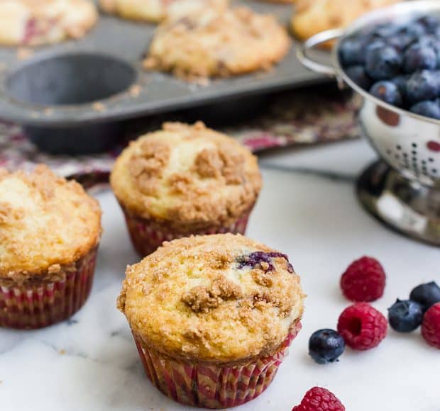 Freshly baked mixed berry muffins with summer berries surrounding them on a countertop.