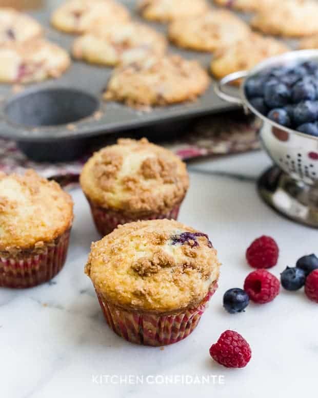 Freshly baked mixed berry muffins with summer berries surrounding them on a countertop.