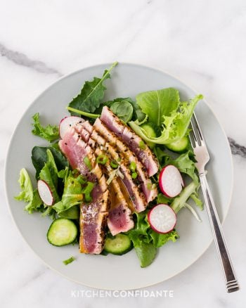 Slices of seared ahi tuna over a bed of mixed greens on a white plate.