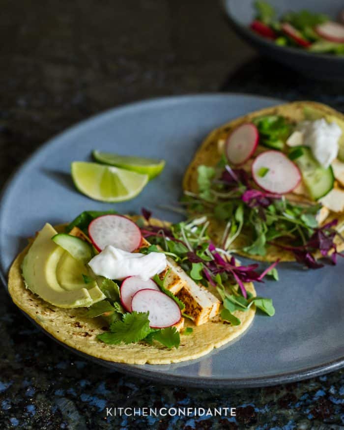 Two tofu tacos on a plate garnished with avocado slices, sliced radishes, sour cream, and micro greens.