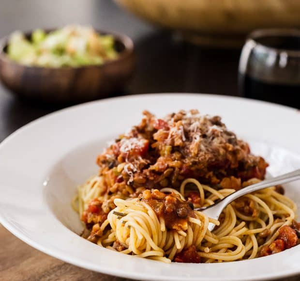 A fork swirled around a bite of Spaghetti with Chicken Sausage and Lentil Bolognese and sauce in a white dish.