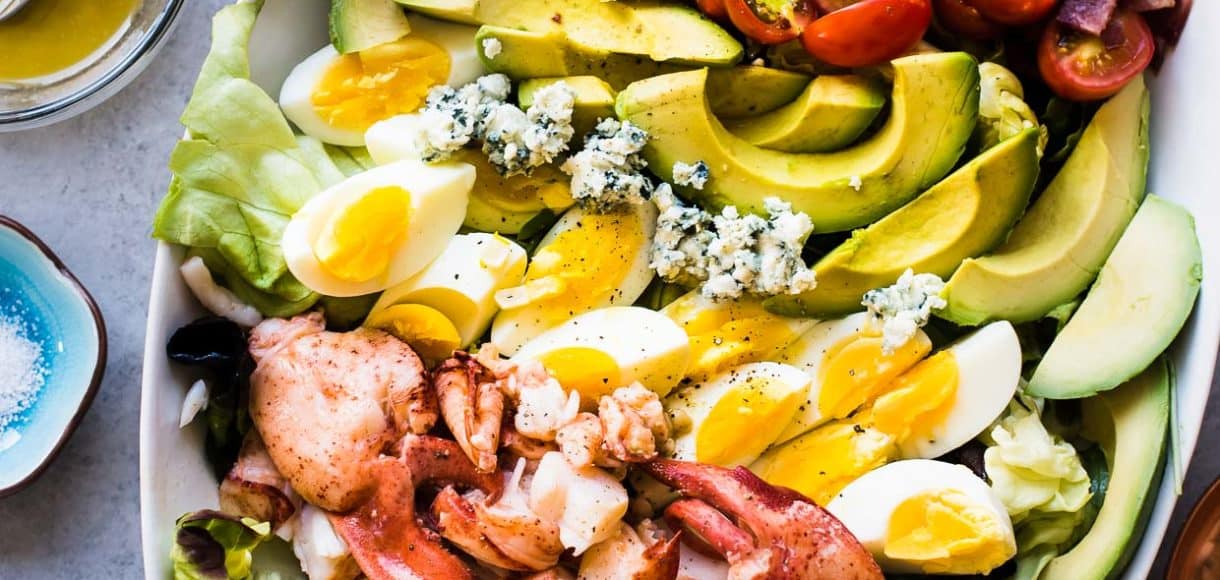 Lobster Cobb Salad on a white platter with lobster, hardboiled eggs, avocados, corn, and fresh greens.