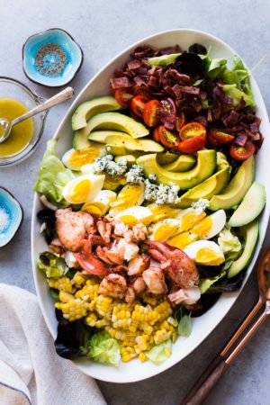 Lobster Cobb Salad on a white platter with lobster, hardboiled eggs, avocados, corn, and fresh greens.