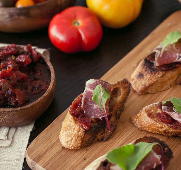 Fire Roasted Tomato Jam Crostini - slices of baguette covered with tomato jam, a slice of salami, and a leaf of baby chard.