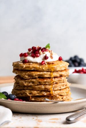 Oatmeal Pancakes stacked on a plate, topped with whipped cream, pomegranate seeds, nuts and maple syrup.