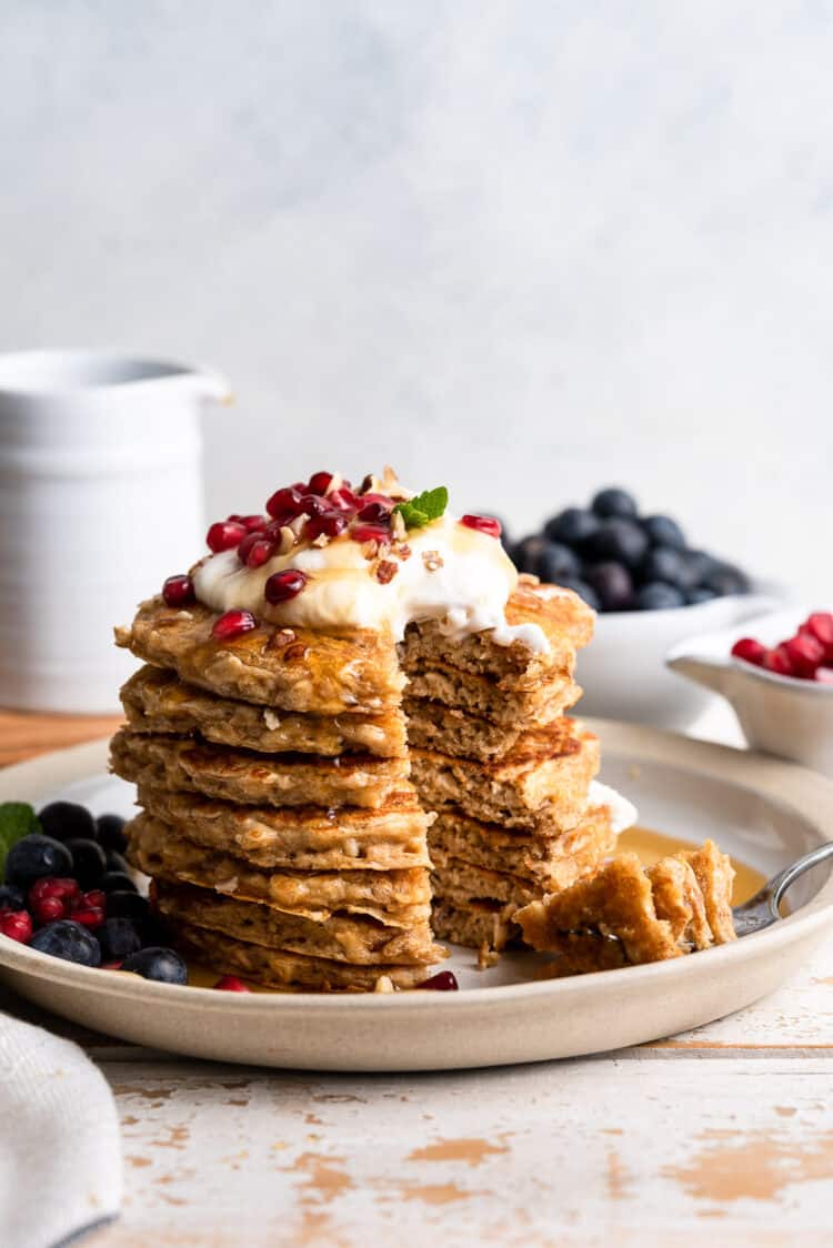 Stack of oatmeal pancakes topped with coconut whipped cream, pomegranate seeds and crushed hazelnuts, and served with maple syrup and blueberries.
