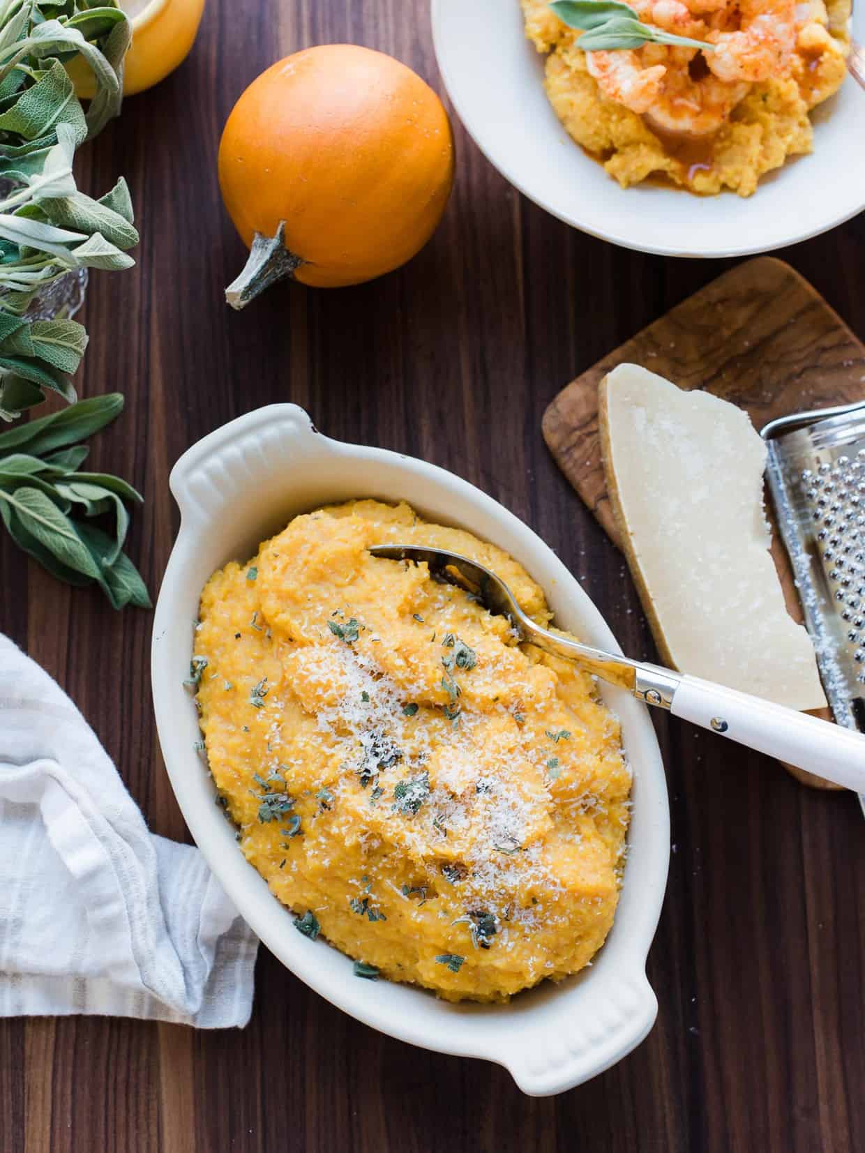 A white serving bowl filled with Pumpkin Sage Polenta surrounded by fresh sage and pumpkins.