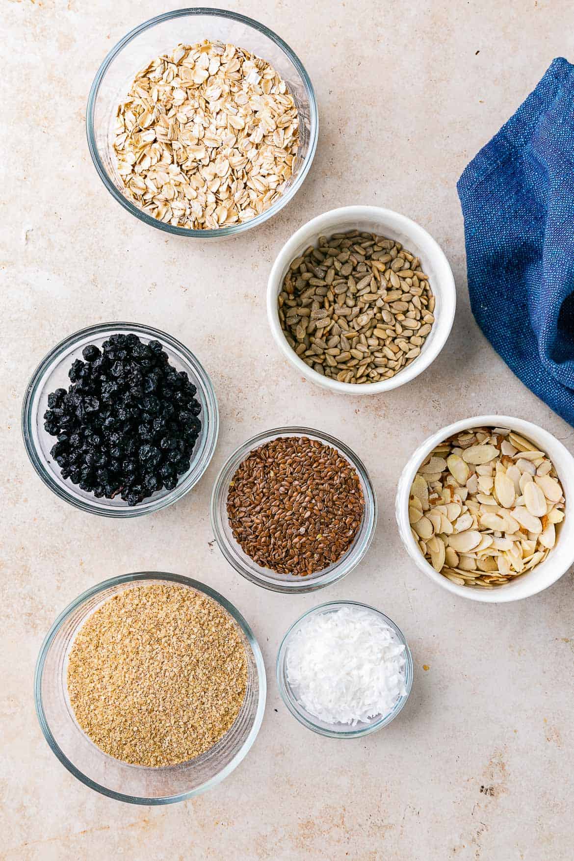 White bowls filled with ingredients to make homemade muesli, like oats, dried blueberries, and flaxseed meal.