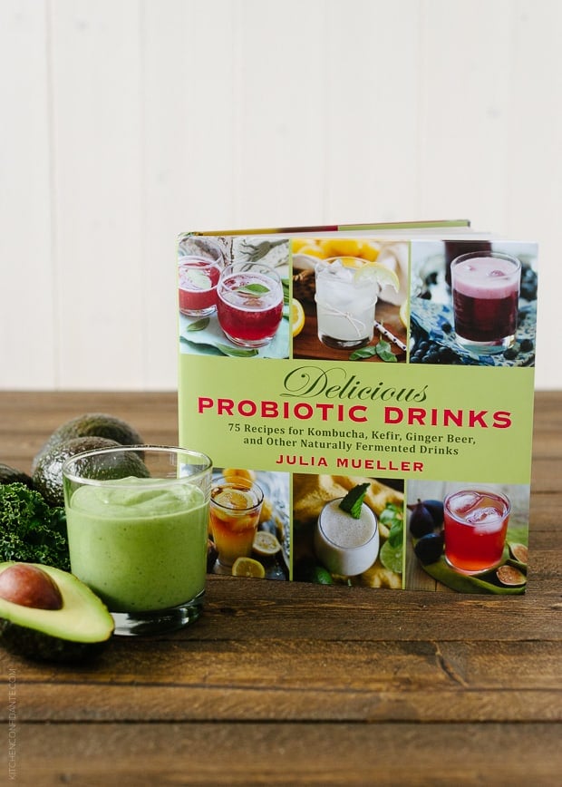 Green Avocado Kale Superfood Smoothie in a glass next to an avocado, kale leaves, and a copy of the Delicious Probiotic Drinks cookbook