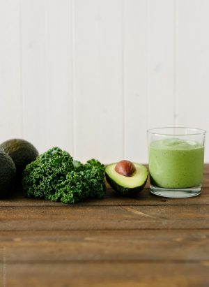 Green Avocado Kale Superfood Smoothie in a tall glass next to an avocado and kale leaves