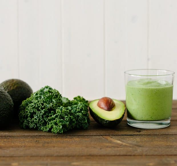 Green Avocado Kale Superfood Smoothie in a tall glass next to an avocado and kale leaves