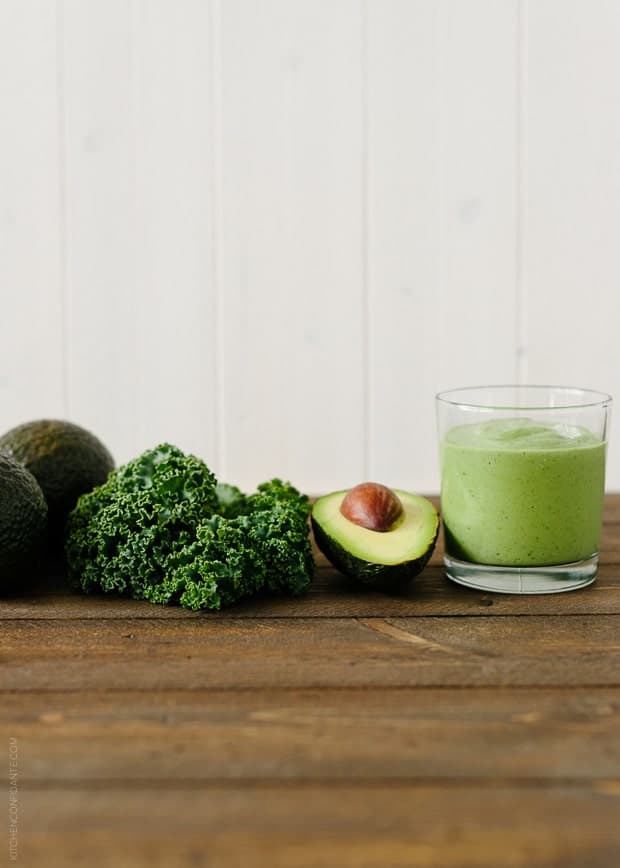 Green Avocado Kale Superfood Smoothie in a tall glass next to an avocado and kale leaves