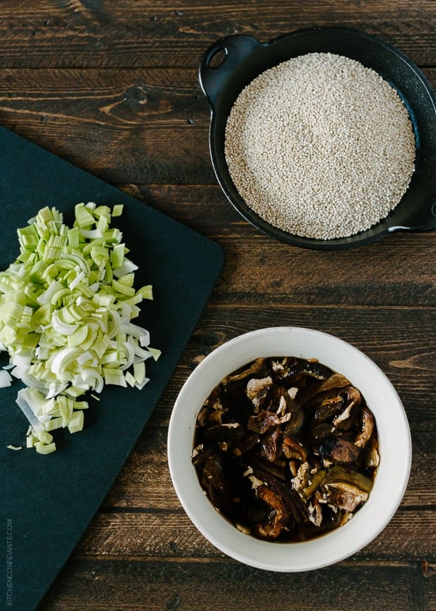 A bowl of quinoa, a bowl of porcini mushrooms, and chopped leeks on a wooden surface.