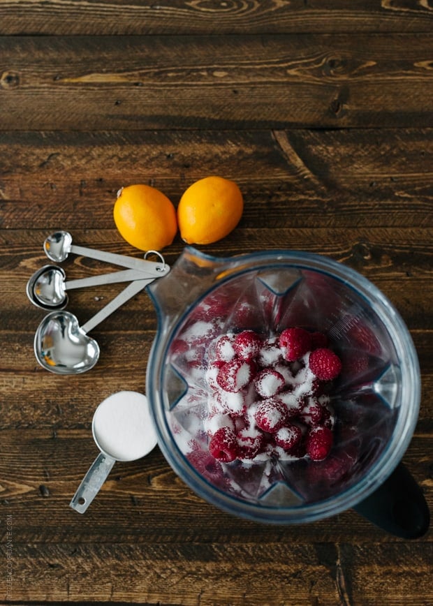 A bowl of fresh raspberries and sugar surrounded by measuring spoons and lemons.