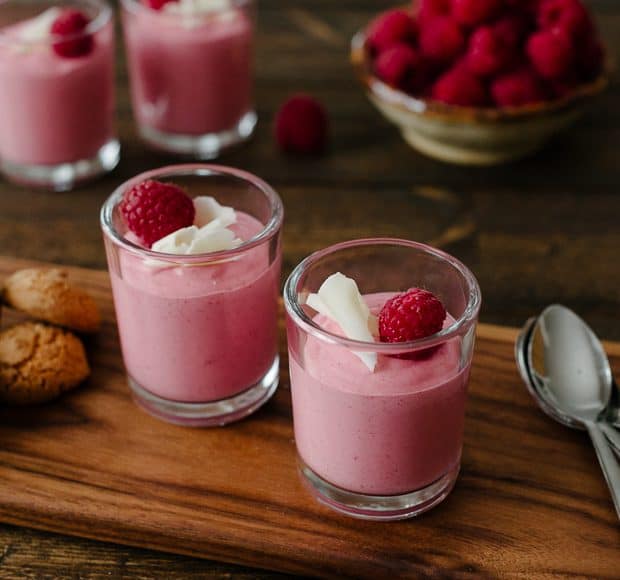 Small glass cups of raspberry mousse garnished with white chocolate and raspberries.