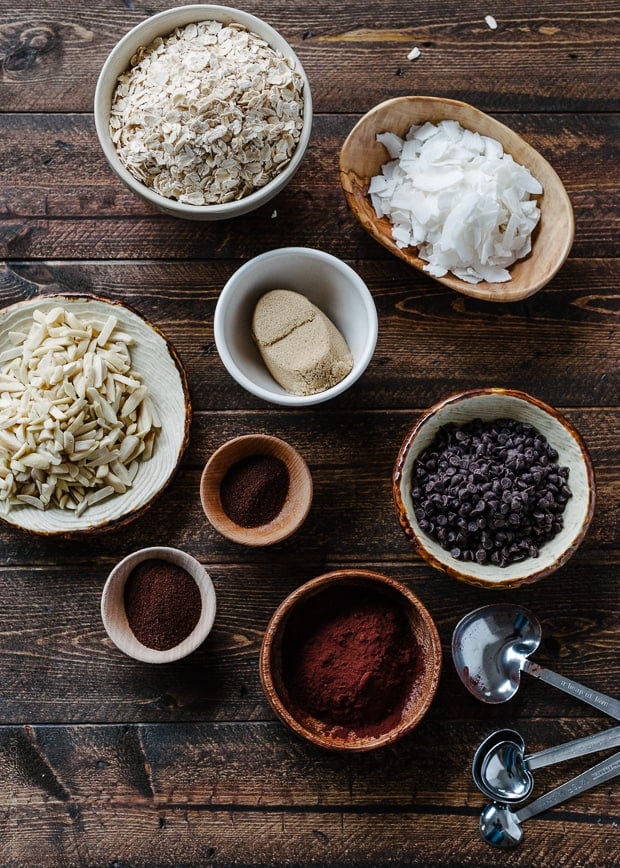 Bowls of oats, slivered almonds, coconut, and more ingredients to make homemade granola arranged on a wooden surface.