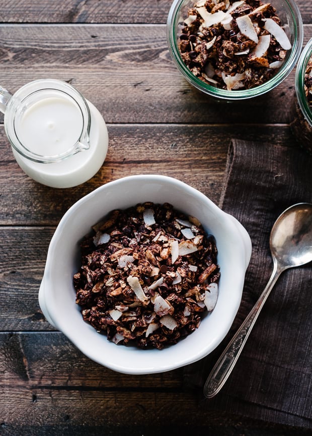 Milk, a spoon, and a bowl of homemade granola on a wooden surface.