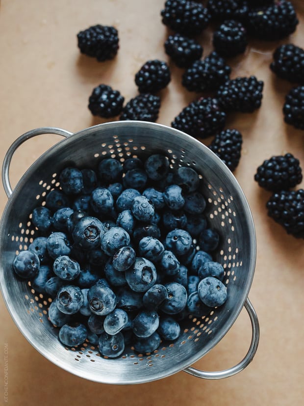 A colander filled with blueberries with blackberries in the background.