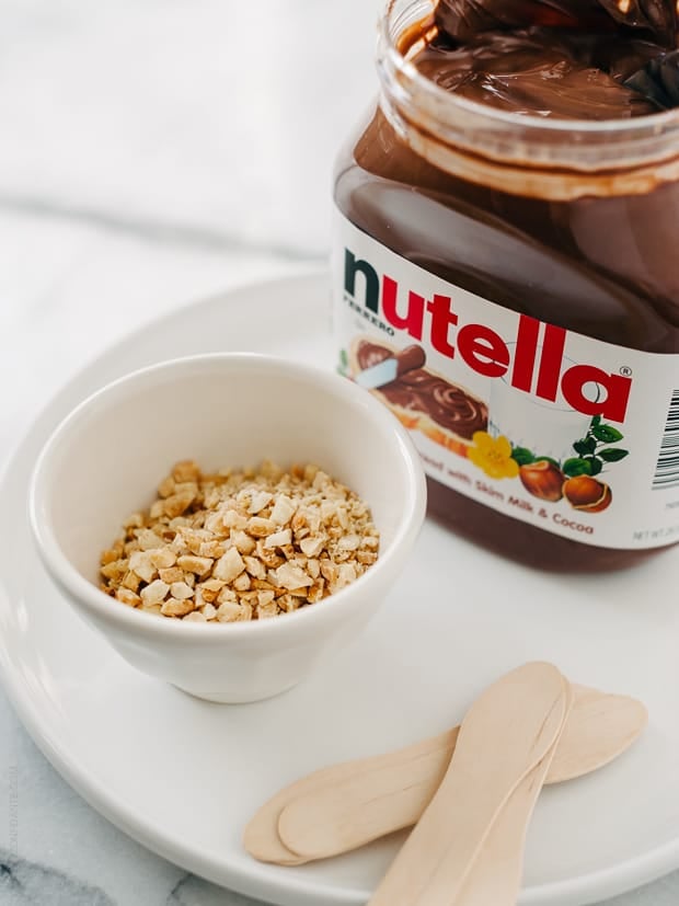 A jar of Nutella, a dish of chopped nuts and popsicle sticks.