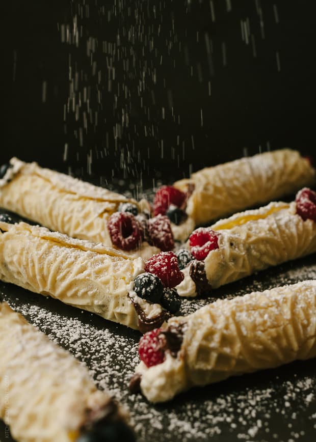 Rolled and filled Pizzelle cookies being sprinkled with powdered sugar.