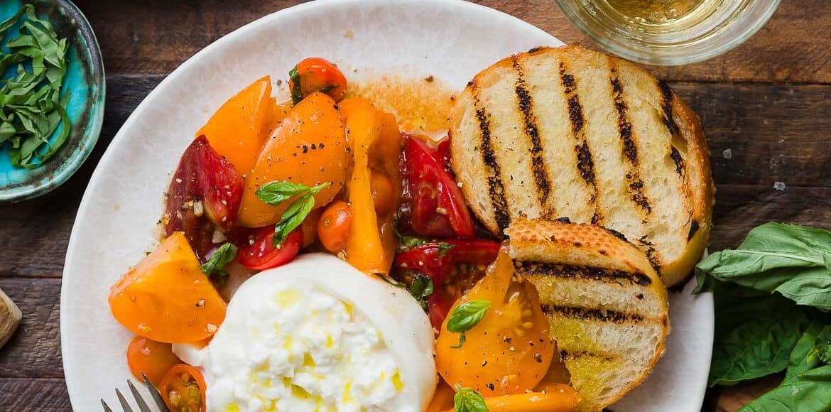 Heirloom tomatoes on a white plate topped with burrata and surrounded with toasted baguette slices.