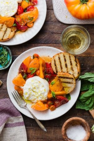 Heirloom tomatoes on a white plate topped with burrata and surrounded with toasted baguette slices.
