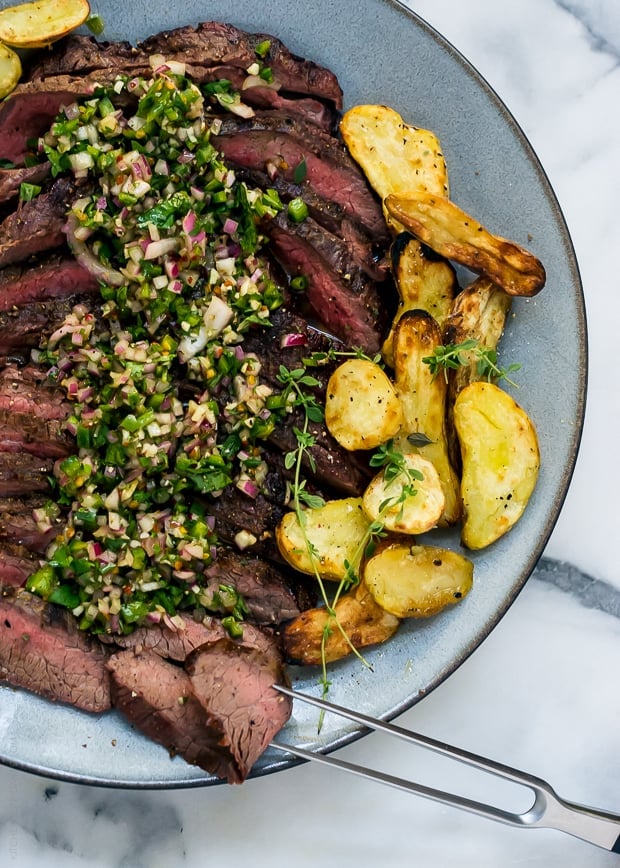 Sliced grilled flank steak surrounded by grilled fingerling potatoes and topped with chimichurri sauce.