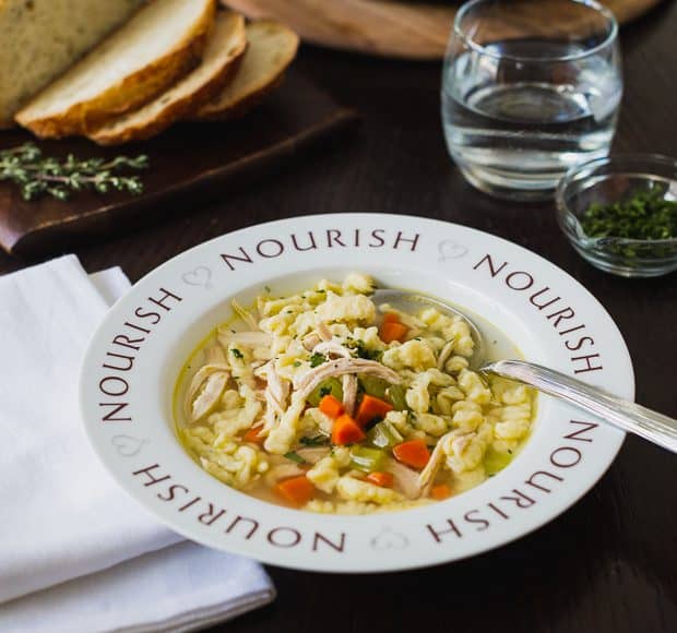 Chicken and Herb Spaetzle Soup served in a bowl with the word, “Nourish” around the rim.