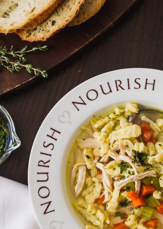 Chicken and Herb Spaetzle Soup served in a bowl with the word, “Nourish” around the rim.