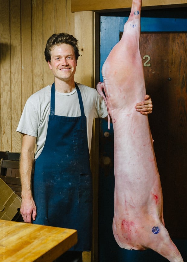 A man standing beside a hanging and recently butchered animal at the Advanced Butchery Class - Avedano's SF. 