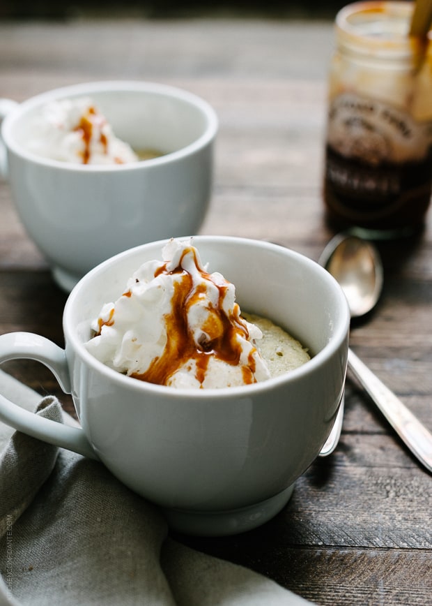 Two banana caramel mug cakes topped with whipped cream on a wooden surface.