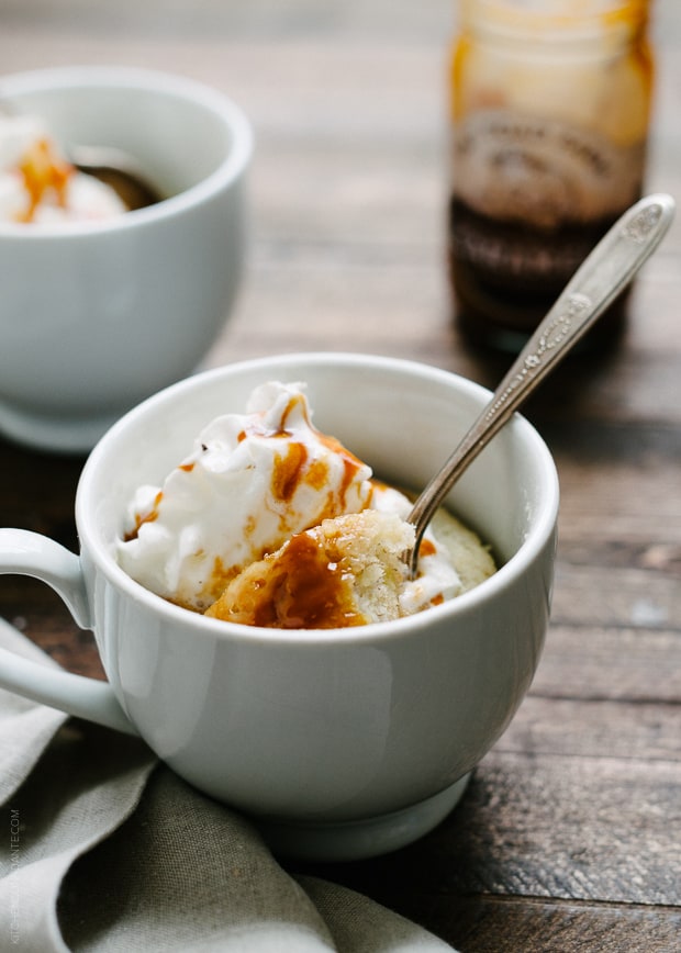 A microwave mug cake topped with caramel and whipped cream on a wooden surface.