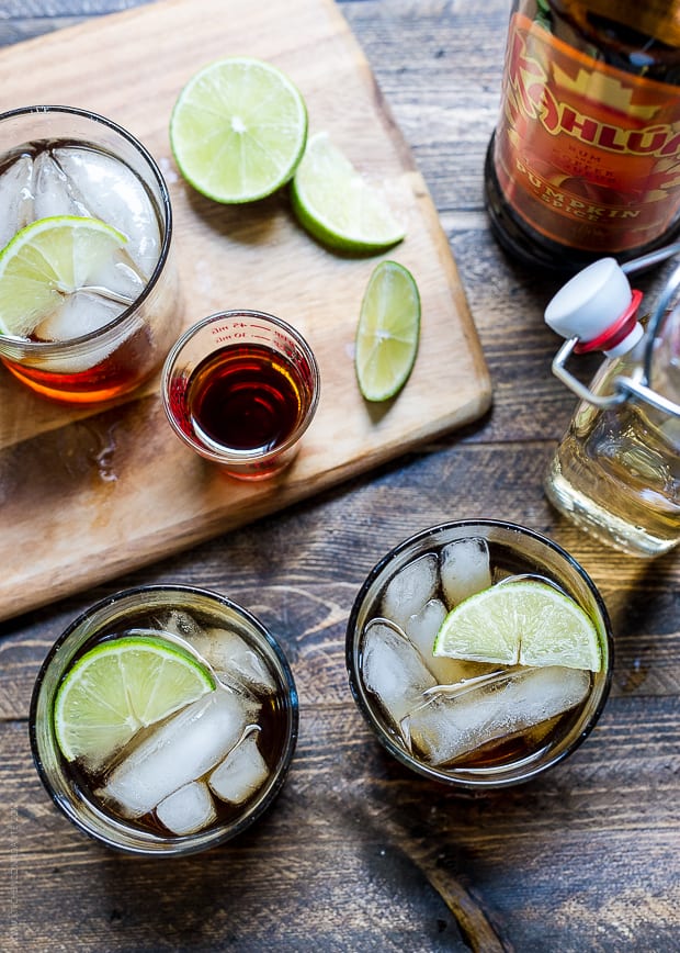 Cocktails on a wooden surface surrounded by a cutting board, slices of lime, ginger beer and Kahlúa Pumpkin Spice.