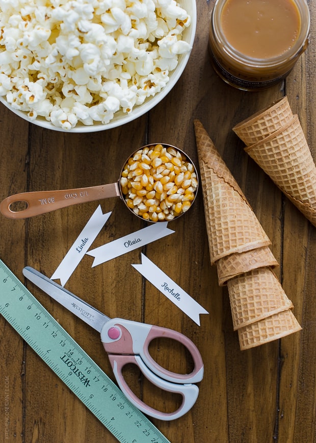 Supplies to make Popcorn Christmas Tree Place Cards including a scoop of popcorn, cardstock, scissors, ice cream cones and caramel.