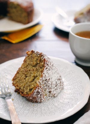 Slice of Banana Pineapple Cake with powdered sugar on a white plate. A cup of tea in the background with banana pineapple cake.