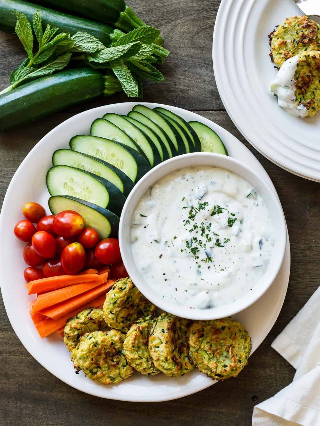 Baked Zucchini Fritters with Ranch Tzatziki on a white plate surrounded by veggies.