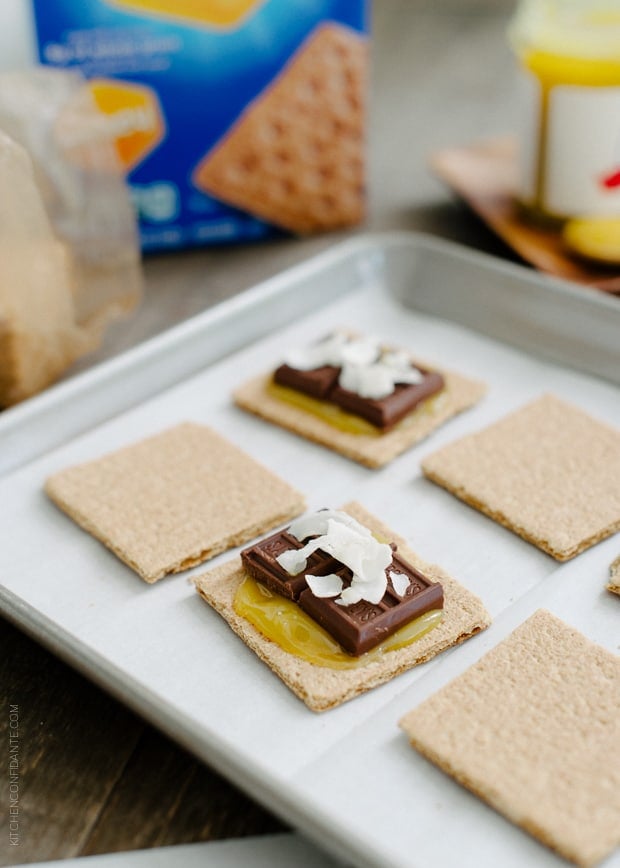 Preparing Coconut Jam Berry S’mores by layering coconut jam, milk chocolate, and shredded coconut on top of graham crackers.