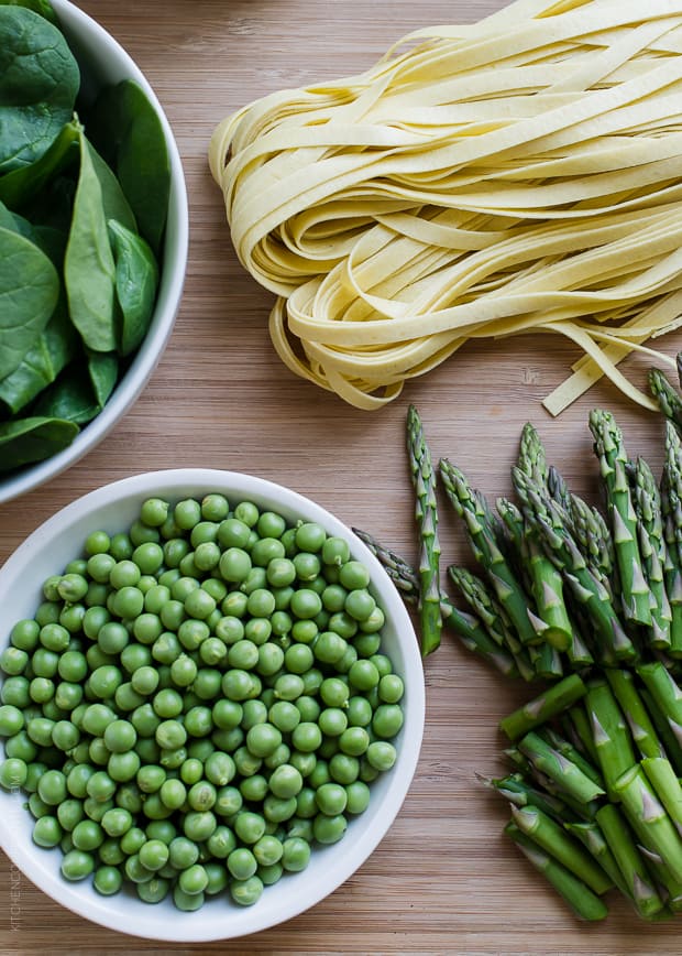 A bowl of spinach, a bowl of peas, chopped asparagus, and fresh fettuccine on a wooden surface.