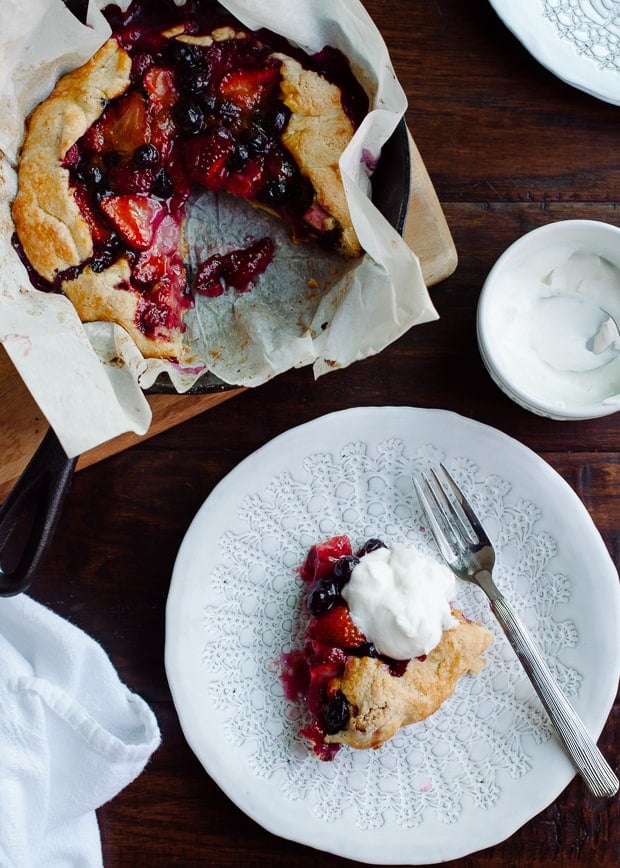 A slice of Mixed Berry Cornmeal Galette on a white plate with a dollop of whipped cream on top.