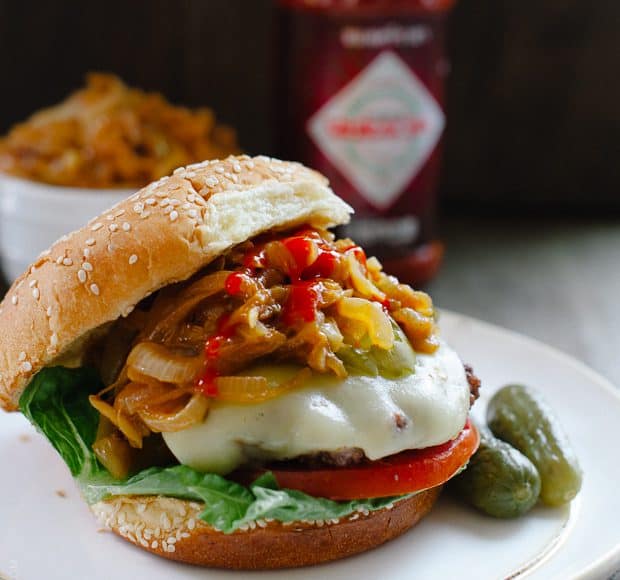 A hamburger loaded with toppings like cheese, tomato, pickles, and Sriracha Caramelized Onions.