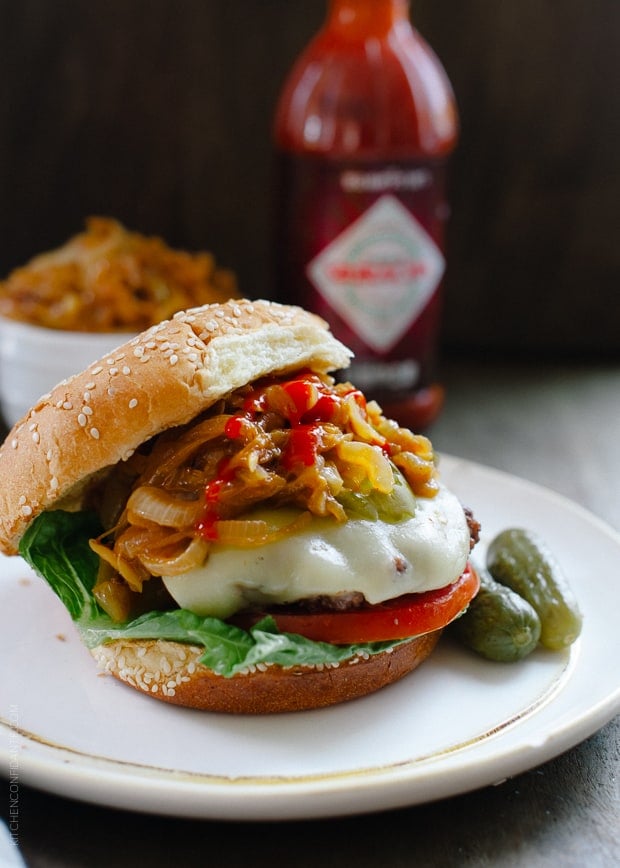 A hamburger loaded with toppings like cheese, tomato, pickles, and Sriracha Caramelized Onions.