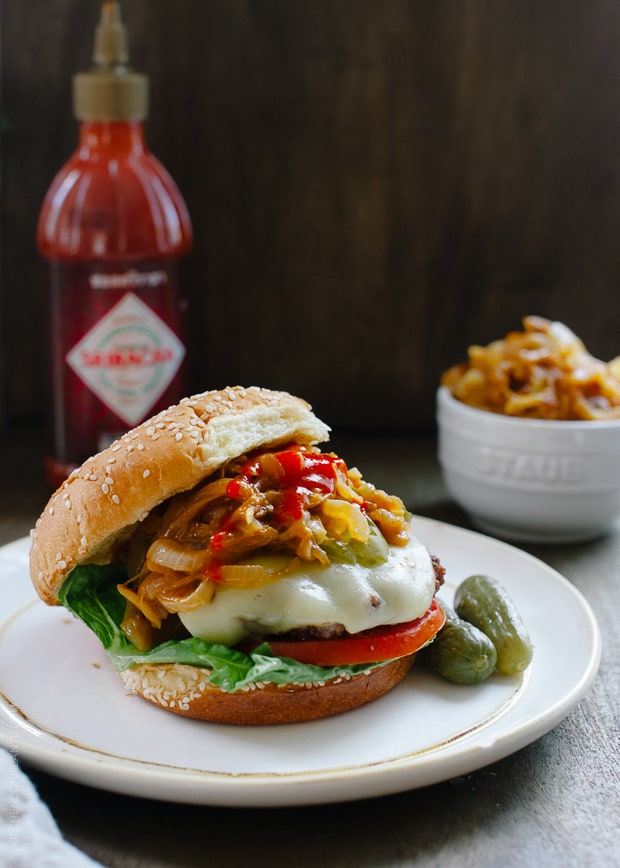 A hamburger loaded with toppings including tomato, lettuce, pickles, cheese, and Sriracha Caramelized Onions. 