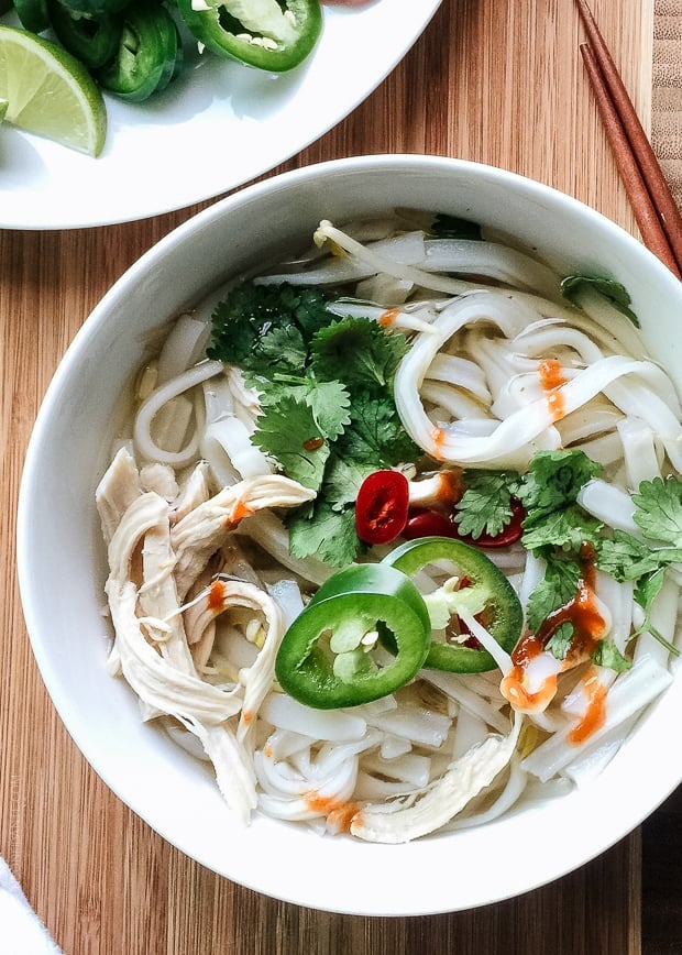 A bowl of easy chicken pho with torn herbs, slices of lime, long chewy noodles, poached chicken and broth.