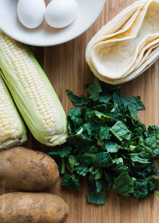Potatoes, corn on the cob, chopped kale, eggs, and taco boat shells on a wooden surface.