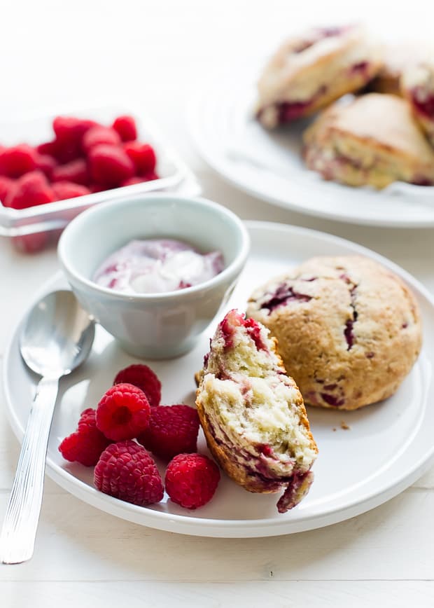 coconut raspberry scones and raspberries on a white plate with coconut raspberry yogurt in a dish, more scones and raspberries in the background