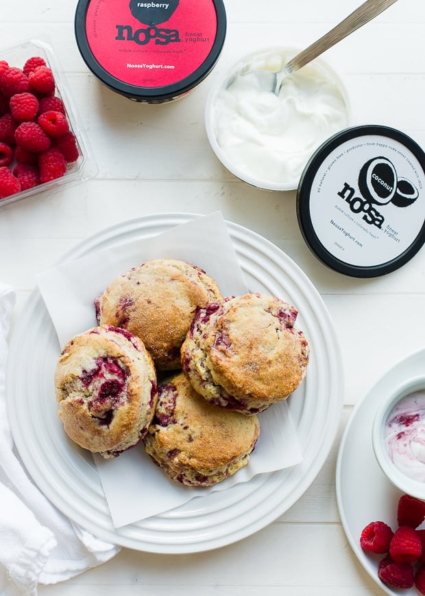 coconut raspberry scones on a white plate surrounded by noosa yogurt containers and raspberries on a white background